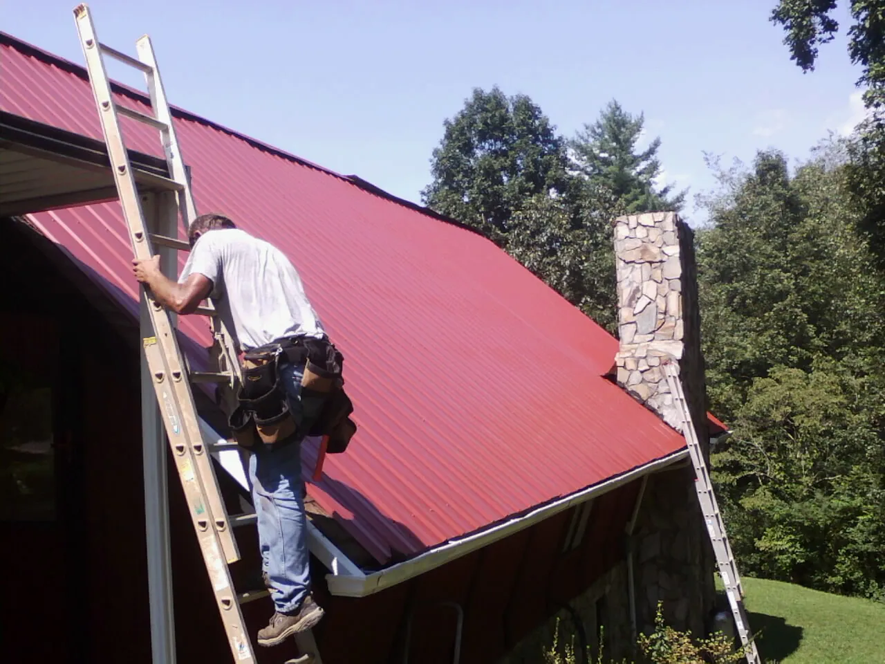 Roofer on a ladder beside a red metal roof and stone chimney — Soffit & Fascia Repair by Roofing Brothers in Corryton